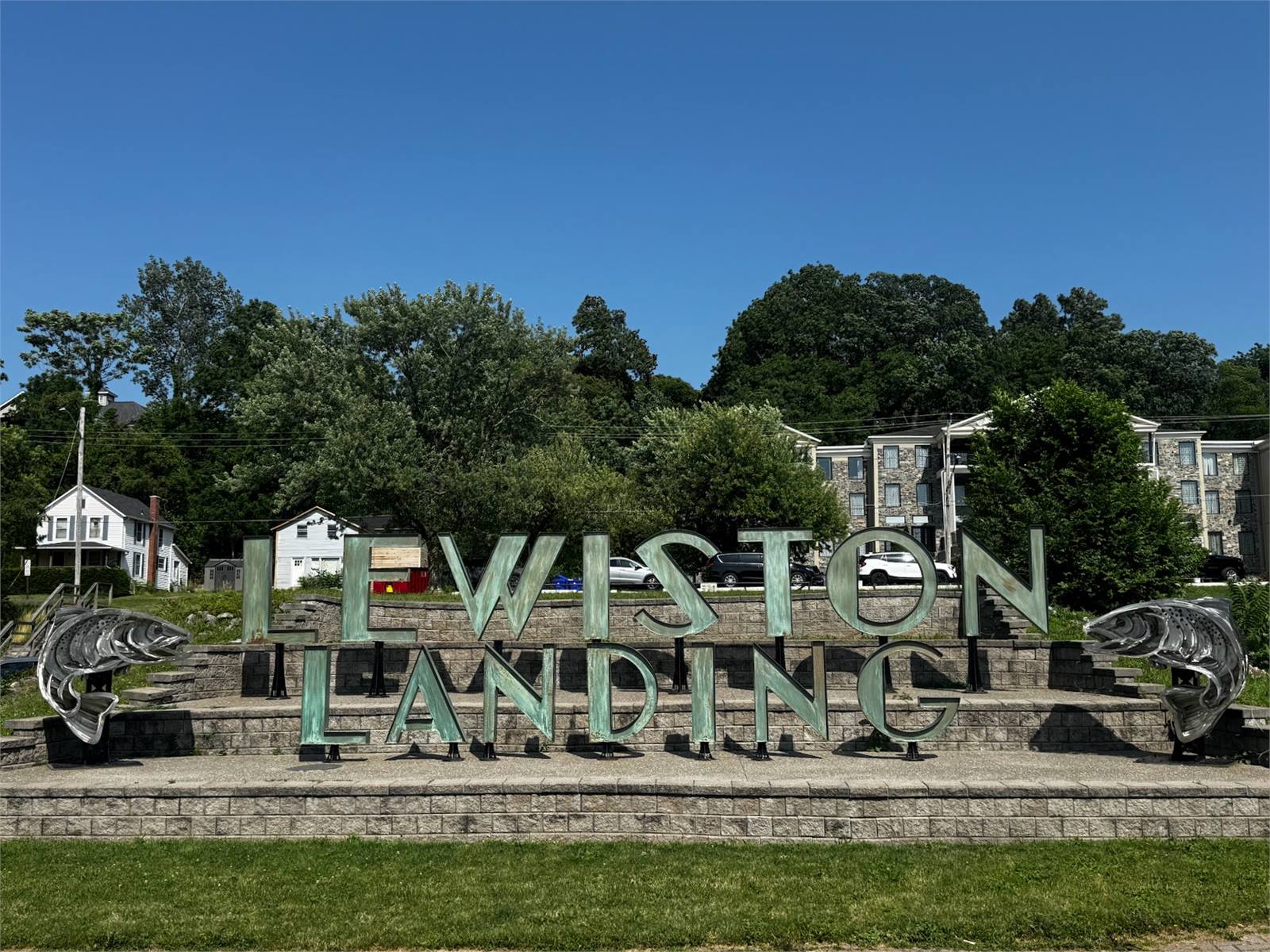 The Lewiston Landing sign on the Niagara River waterfront, flanked by two steel salmon sculptures under a clear blue sky.