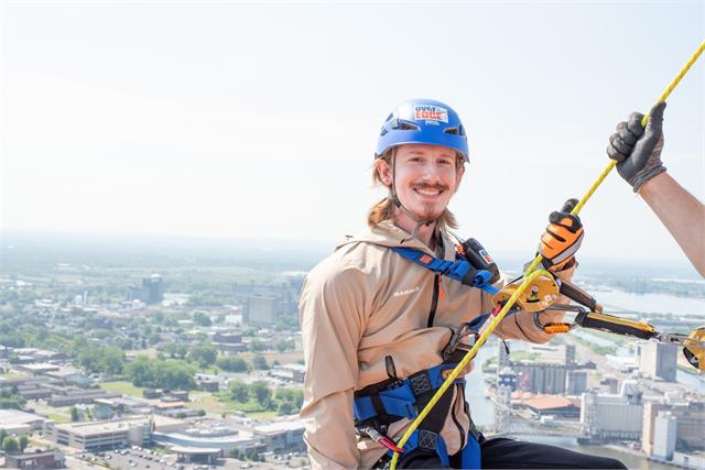 Garrett Lester in a helmet and harness rappelling down the Seneca One tower during the Over the Edge charity fundraiser, with Buffalo spread out far below.